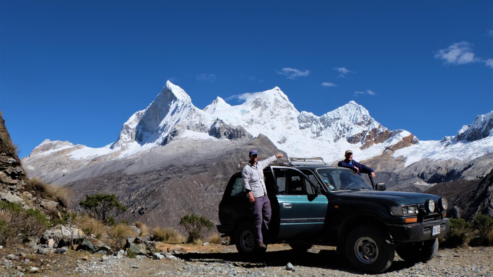 Two men standing on a car in front of a snowcapped Peruvian mountain
